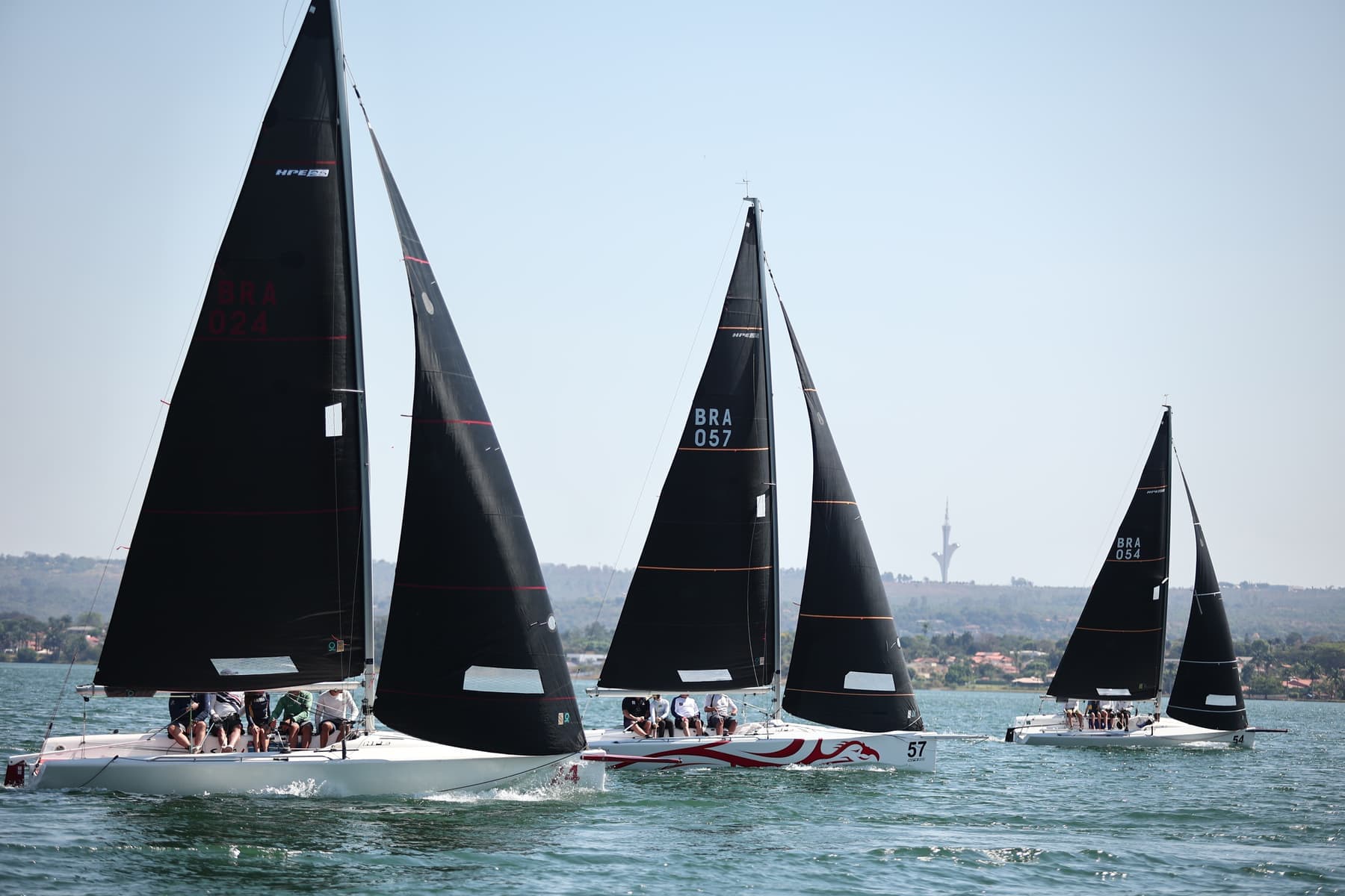 Frota de barcos de quilha com velas escuras navegando lado a lado no Lago Paranoá, com a orla de Brasília ao fundo